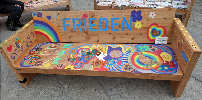Bemalte Holzbank mit FRIEDEN und Friedenssymbolen wie den Regenbogen, Taube...